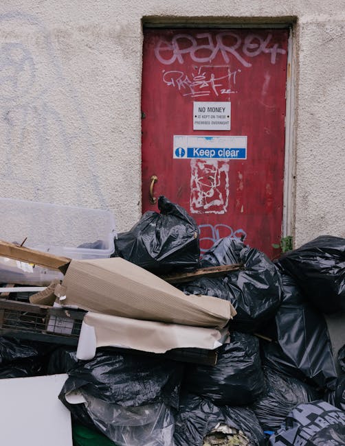 A pile of black garbage bags filled with waste materials, along with discarded cardboard, packaging foam, and wooden debris, is stacked in front of a red security door with graffiti tags and multiple warning signs including 'No Money' and 'Keep Clear.' The scene is outdoors near a building wall, and the waste appears to be ready for disposal or collection, reflecting proper containerization in a home relocation or moving process supported by Removal Companies Marylebone. The area is illuminated by natural light, with the bags and materials placed on the pavement, indicating clearing up after packing or unloading during a house move or furniture transport service.