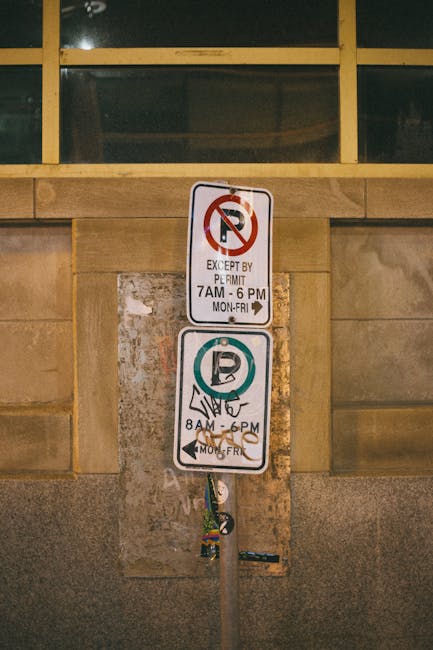 A vertical metal post with two rectangular parking restriction signs mounted one above the other, positioned against a stone wall beneath a window with multiple panes. The top sign indicates no parking except by permit from 7 am to 6 pm, Monday to Friday, with a red circle and diagonal line over a black P symbol, and a white background. The bottom sign shows a parking symbol within a circle, with a diagonal line through it, indicating no parking from 8 am to 6 pm, Monday to Friday, also on a white background. Behind the signs, the wall features large rectangular stones in a beige-brown tone with a slightly textured surface, and the window above has a grid of black frames and glass panes. In the context of home relocation and furniture transport by Removal Companies Marylebone, these signs highlight parking restrictions relevant to loading and unloading processes during house removals or moving services.