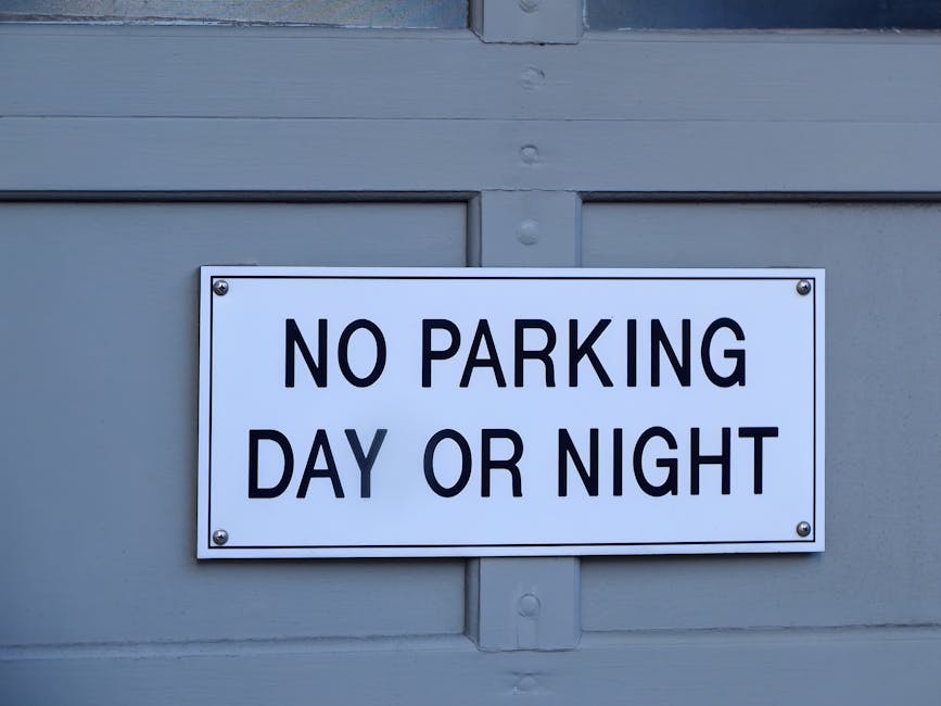 A vertical metal post with two rectangular parking restriction signs mounted one above the other, positioned against a stone wall beneath a window with multiple panes. The top sign indicates no parking except by permit from 7 am to 6 pm, Monday to Friday, with a red circle and diagonal line over a black P symbol, and a white background. The bottom sign shows a parking symbol within a circle, with a diagonal line through it, indicating no parking from 8 am to 6 pm, Monday to Friday, also on a white background. Behind the signs, the wall features large rectangular stones in a beige-brown tone with a slightly textured surface, and the window above has a grid of black frames and glass panes. In the context of home relocation and furniture transport by Removal Companies Marylebone, these signs highlight parking restrictions relevant to loading and unloading processes during house removals or moving services.