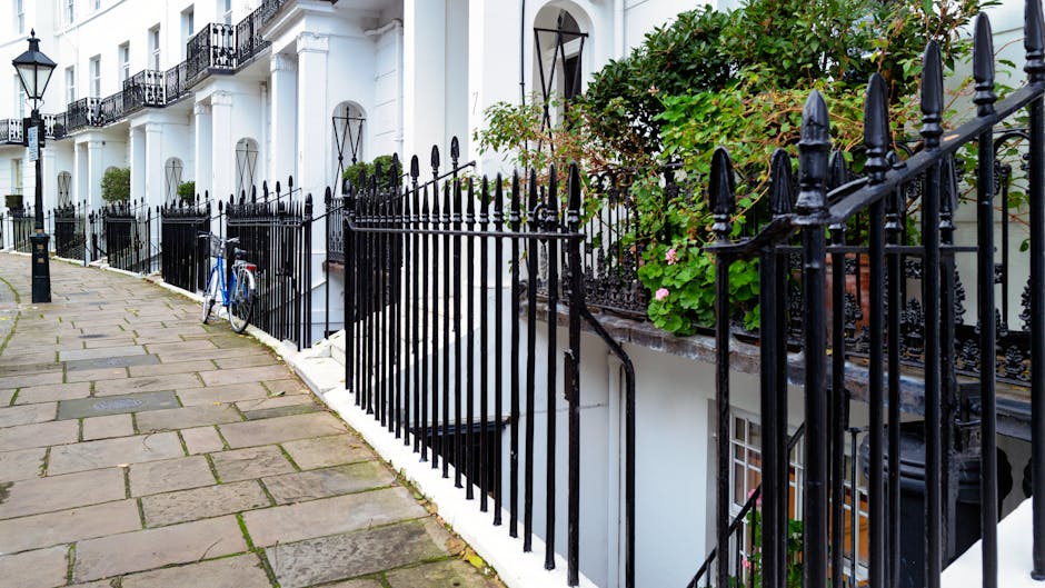 Image showing the exterior of a white Victorian-style terraced house on Baker Street, Marylebone, with a black wrought iron fence along the pavement. The sidewalk is paved with large rectangular stone slabs, and a blue bicycle is parked against the fence. The house has tall windows with decorative black ironwork above the window sills, and some greenery including flowering plants in pots and window boxes is visible along the front. The outdoor scene is well-lit, and the photograph captures prepared house removal activities relevant to local relocation services provided by Removal Companies Marylebone, including moving logistics and home furniture transport in narrow terraced streets.