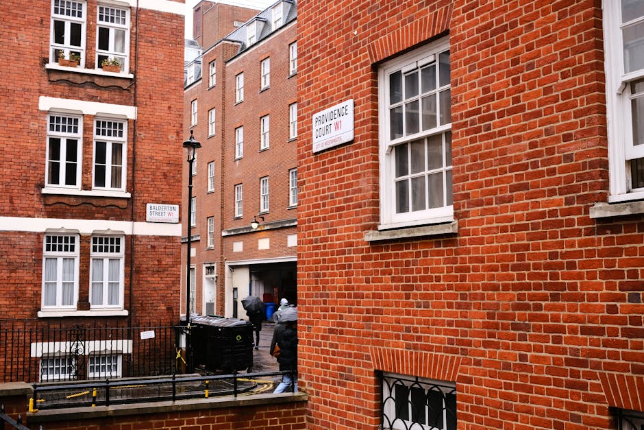 The image shows a narrow street in Marylebone with red brick residential buildings. Two workers from Removal Companies Marylebone are visible, dressed in grey jackets, carrying a large piece of furniture wrapped in plastic and secured with straps. They are positioned near the entrance of a building, navigating a cluttered area with black waste bins and a trolley nearby. The sidewalk leading up to the entrance is partially obstructed, indicating an active home relocation process involving furniture transport through tight terraces. The scene is lit with natural daylight, capturing the urban environment and the logistical challenges of moving large items in constrained spaces as part of professional removals service.