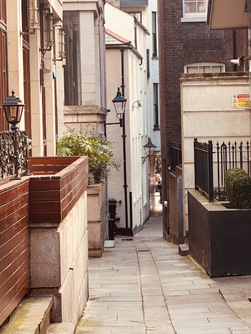 Image showing the exterior of a white Victorian-style terraced house on Baker Street, Marylebone, with a black wrought iron fence along the pavement. The sidewalk is paved with large rectangular stone slabs, and a blue bicycle is parked against the fence. The house has tall windows with decorative black ironwork above the window sills, and some greenery including flowering plants in pots and window boxes is visible along the front. The outdoor scene is well-lit, and the photograph captures prepared house removal activities relevant to local relocation services provided by Removal Companies Marylebone, including moving logistics and home furniture transport in narrow terraced streets.