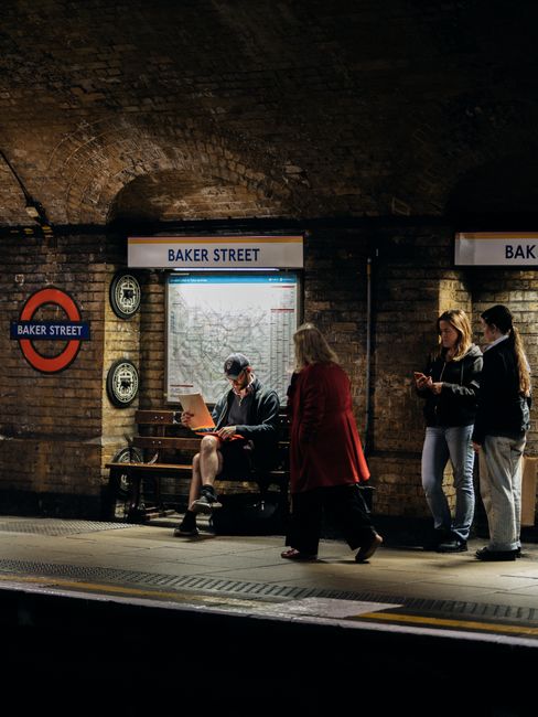 A man sitting on a wooden bench at Baker Street underground station, engrossed in reading a document while wearing a blue cap and glasses, with a woman in a red coat standing nearby and two other women with light-colored hair and dark jackets waiting in line. The station features exposed brick walls, a blue and white station sign reading 'BAKER STREET', and vintage-style telephone booths mounted on the wall. The lighting highlights the textured brickwork and creates a subdued atmosphere. This scene captures the typical interior of a London underground station, relevant to house removals and moving logistics by illustrating the transit environment associated with furniture transport and home relocation.