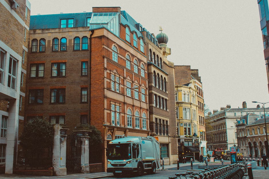 The image depicts a city street scene during daytime in Marylebone, featuring a mix of historic and modern multi-storey buildings with brick and light-colored facades. In the foreground, a light blue removal vehicle from Removal Companies Marylebone is parked on the paved street, with the side of the van visible. Nearby, a trolley or moving dolly is loaded with large cardboard boxes and wrapped furniture, likely part of a home relocation process. Several packed cardboard boxes, some wrapped in plastic for protection, are visible on the pavement beside the vehicle, indicating ongoing packing or loading activities. The vehicle is positioned close to an entrance of a property, with some boxes being carried inside or prepared for transport. The street has a row of metal bike racks, and background buildings include shops and offices, with overcast lighting giving a neutral tone to the scene, illustrating typical furniture transport and packing and moving operations supported by [COMPANY_NAME] within the context of house removals and relocation services.
