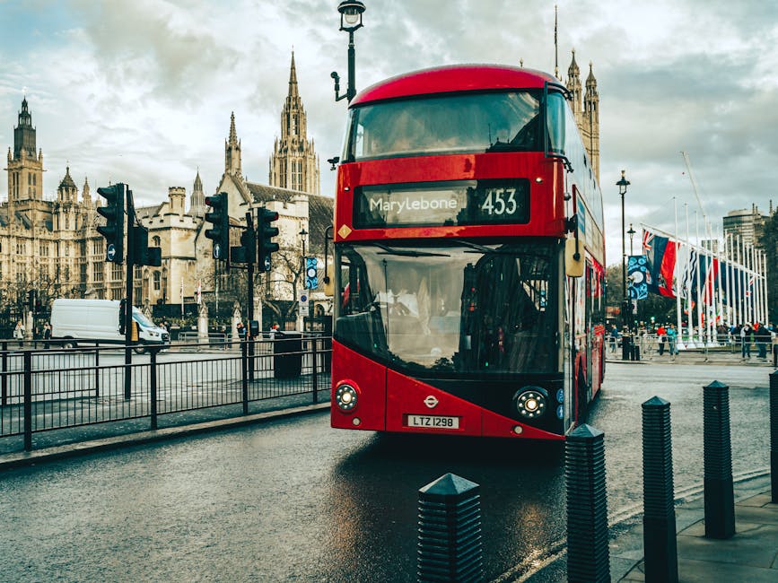 A red double-decker bus displaying the route number '453' and the destination 'Marylebone' is parked on a wet street in front of a historic architectural background featuring a large Gothic-style building with multiple spires. The bus is part of the London bus service, with the license plate 'LTZ 1298'. Nearby, a white van is visible driving along the road, and several pedestrians are walking on the pavement. Traffic lights and street lamps are mounted along the road, with some green lights illuminated. The overcast sky indicates rainy or gloomy weather, and reflections of the bus and surroundings can be seen on the damp pavement. This scene showcases typical urban transport infrastructure, which Removal Companies Marylebone might consider when planning home relocation logistics involving furniture transport and loading processes within the city.