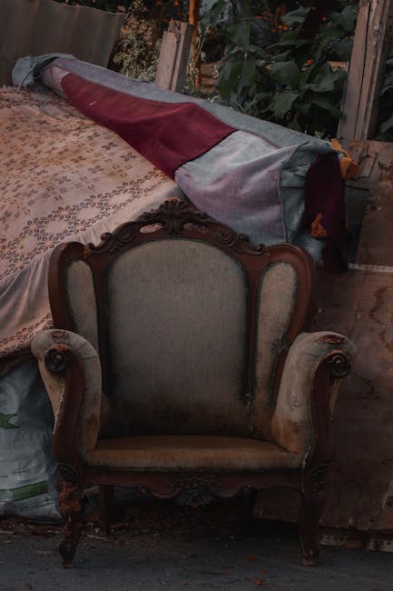 A vintage armchair with ornate wooden carvings and beige upholstery is situated indoors next to a large piece of furniture covered with a multicolored protective moving blanket, secured with straps. The armchair is positioned on a wooden floor, with a floral-patterned rug partially visible beneath it. In the background, there are green plants and a wooden structure, indicating a rustic environment. The scene suggests a home relocation or furniture transport process, with the furnishings prepared for moving or packing, as handled by Removal Companies Marylebone during a house removal or furniture transport operation.