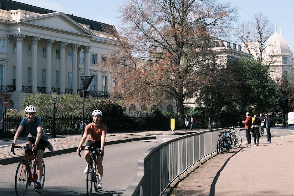 Two cyclists wearing helmets riding along a street with historic white buildings featuring columns and decorative facades, while several pedestrians walk on the sidewalk. On the right side, there is a metallic railing with bicycles locked to it, and a group of people standing nearby. The scene includes evenly spaced leafless trees, some with budding leaves, and a large domed building visible in the background under clear skies. The environment suggests a city area, suitable for house removals and relocation logistics, as observed in professional moving services by Removal Companies Marylebone. The street is illuminated by natural daylight, with shadows cast by the trees and other objects, capturing a typical busy urban setting for home relocation or furniture transport activities.