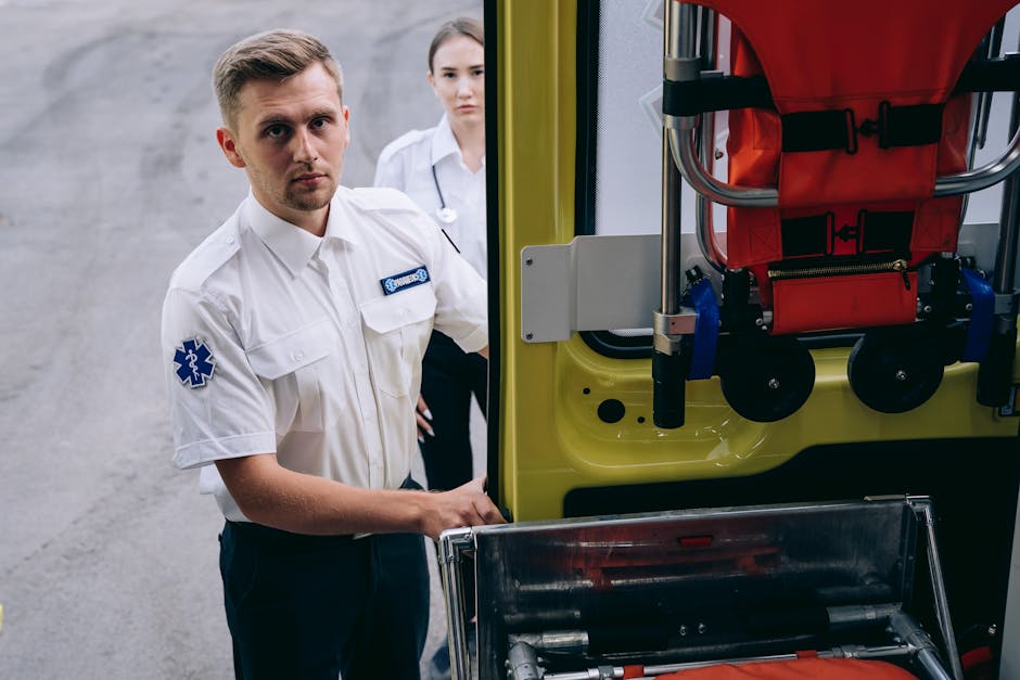 A male paramedic in a white uniform with a blue medical insignia on his sleeve stands beside an open emergency vehicle, which is painted in bright yellow and equipped with a red collapsible wheelchair secured inside. A female medical professional in a white coat is visible in the background, partially obscured, inside the same setting. The scene is indoors, with a plain gray wall and concrete floor, and the paramedic appears to be preparing or inspecting equipment for a home relocation involving urgent medical transport. The image focuses on the loading process of emergency medical equipment, highlighting safety straps, wheelchair, and the vehicle’s interior, reflecting aspects of emergency service and rapid response related to medical evacuations or urgent relocations.