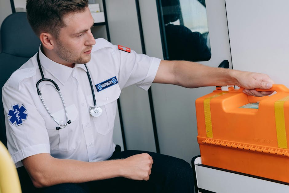 A male emergency medical professional with light skin and short brown hair, wearing a white uniform with patches on the sleeves and a stethoscope around his neck, is seated inside an ambulance near an open window. He is carefully holding a large orange emergency medical kit secured with yellow reflective tape, poised to transport it through the vehicle. The ambulance’s interior features a white wall and storage compartments, with natural light illuminating the scene. This image captures the preparation and handling of emergency medical supplies, relevant to house removals and logistics services like those offered by Removal Companies Marylebone, especially during urgent or time-sensitive relocation scenarios involving careful packing and transport.