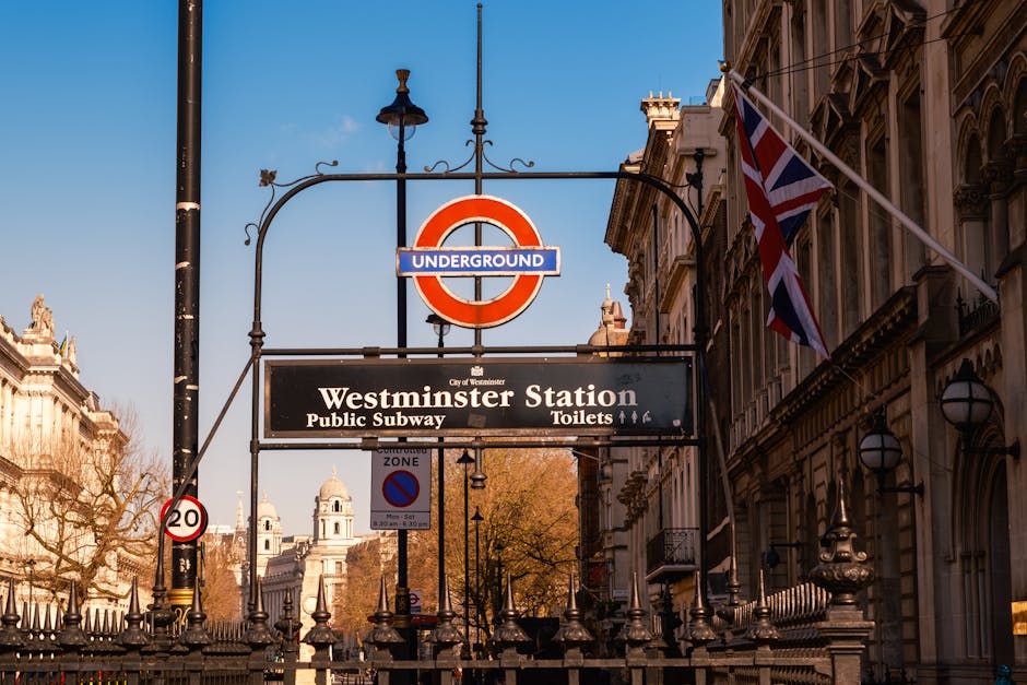 Photograph of Westminster Station in Marylebone, featuring the iconic London Underground roundel sign with a red circle and blue bar displaying the station name. Beneath the roundel, there is a black sign with white text indicating amenities such as public subway and toilets, often used during home relocation or furniture transport processes. The station is set against a backdrop of historic buildings with ornate facades, and Union Jack flags are visible on the right side of the image, fluttering in the breeze. A traditional black lamppost with a decorative design is positioned to the left of the signage. The scene takes place during daylight, with clear blue skies and warm sunlight illuminating the setting. This environment reflects the typical urban landscape encountered during house removals or moving services in Westminster, where logistical planning often involves navigating busy city streets with moving equipment, carrying furniture, or parking removal vans nearby for loading and unloading within the visible vicinity.