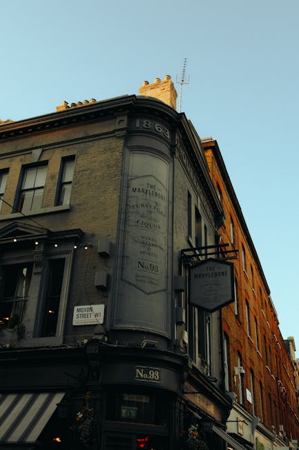 A three-story building located on a street corner in Marylebone, featuring a prominent sign reading 'The Marylebone' and a historical plaque indicating its establishment in 1865. The facade is painted in muted grey and ochre tones, with large windows on each floor, some of which are open. The ground level has a storefront with an awning and a small sign for 'Marylebone' visible near the entrance. The building's roofline has decorative stonework, and a chimney protrudes at the top. The street corner includes a traffic sign and a small potted plant near the doorway. The sky above is clear with a subtle blue hue, and soft natural lighting highlights the building's details, suggesting it is late afternoon. This setting may relate to a home relocation or furniture transport context involving professional removals, as implied by the exterior of the historic retail and service building associated with Removal Companies Marylebone.