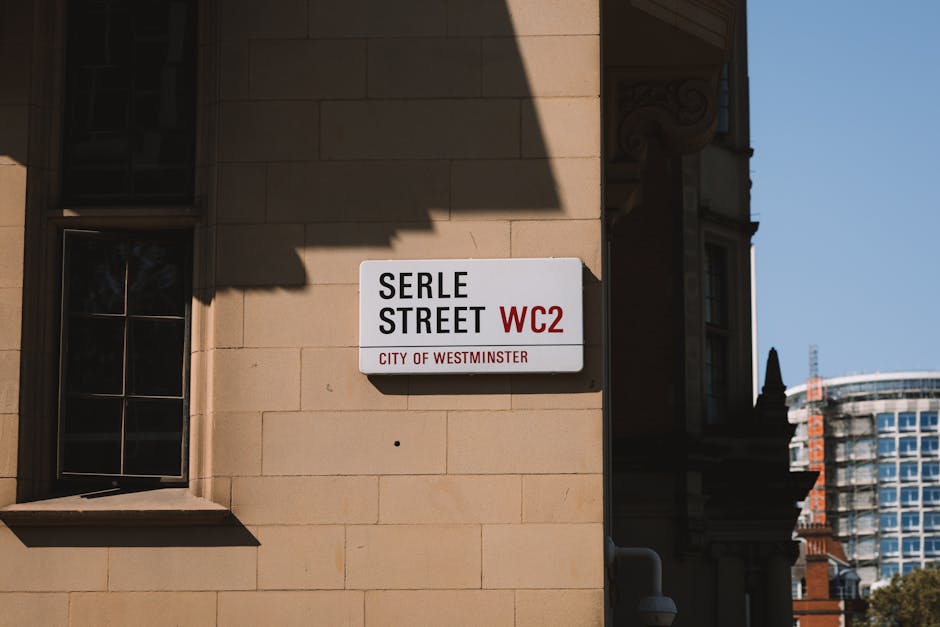 Photograph of Westminster Station in Marylebone, featuring the iconic London Underground roundel sign with a red circle and blue bar displaying the station name. Beneath the roundel, there is a black sign with white text indicating amenities such as public subway and toilets, often used during home relocation or furniture transport processes. The station is set against a backdrop of historic buildings with ornate facades, and Union Jack flags are visible on the right side of the image, fluttering in the breeze. A traditional black lamppost with a decorative design is positioned to the left of the signage. The scene takes place during daylight, with clear blue skies and warm sunlight illuminating the setting. This environment reflects the typical urban landscape encountered during house removals or moving services in Westminster, where logistical planning often involves navigating busy city streets with moving equipment, carrying furniture, or parking removal vans nearby for loading and unloading within the visible vicinity.