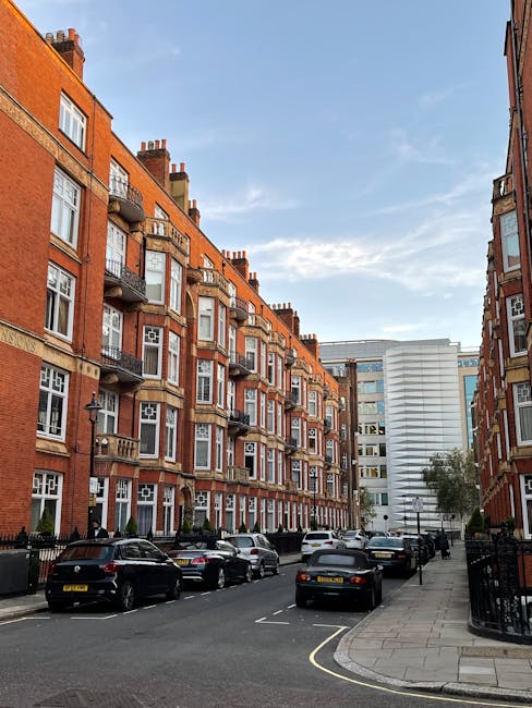 A residential street scene featuring a row of red-brick apartment buildings with multiple floors, each with white-framed windows and small balconies with black metal railings. Several parked cars, including sedans and hatchbacks, line the street on both sides, some with visible license plates. The pavement is wide, with a concrete sidewalk on the right side, and a black metal fence bordering the properties. In the foreground, a loading area is visible where furniture, cardboard boxes, and packing materials are being prepared for home relocation. The sky is partly cloudy with patches of blue, and there are trees and other modern buildings in the background. The image provides a clear view of urban moving logistics, where [COMPANY_NAME] might be involved in furniture transport and packing during a house removal on Wimpole Street and Cavendish Square.